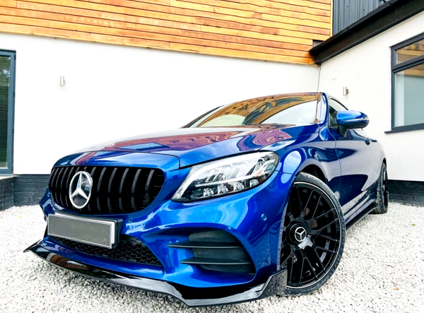 A blue Mercedes-Benz car is parked on gravel, featuring glossy black wheels and a prominent front grille, situated in front of a modern building with white walls and wooden accents.