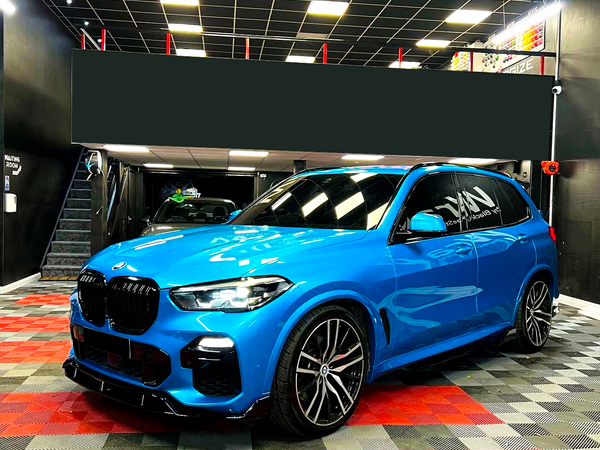 A blue BMW SUV is parked in a garage with a black ceiling and checkered red and black floor. "NAV" is written on the driver's side window.