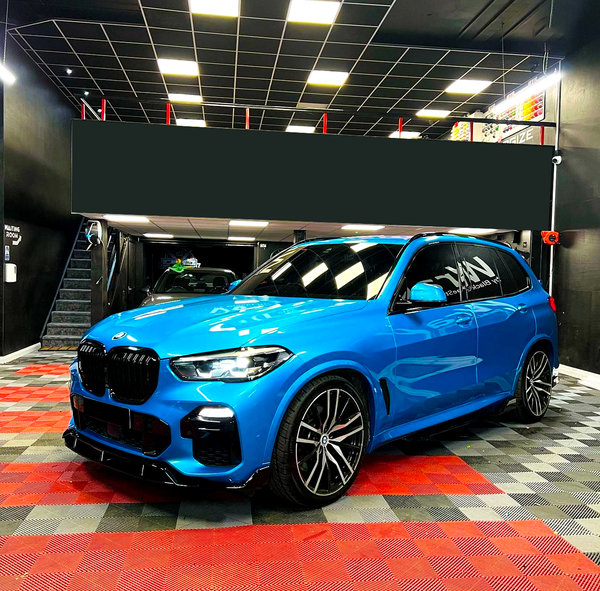 A blue BMW SUV is parked in a garage with a black ceiling and checkered red and black floor. "NAV" is written on the driver's side window.