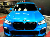 A blue BMW SUV is parked with headlights on in a showroom, surrounded by various wheels displayed on the wall and a checkered floor pattern.