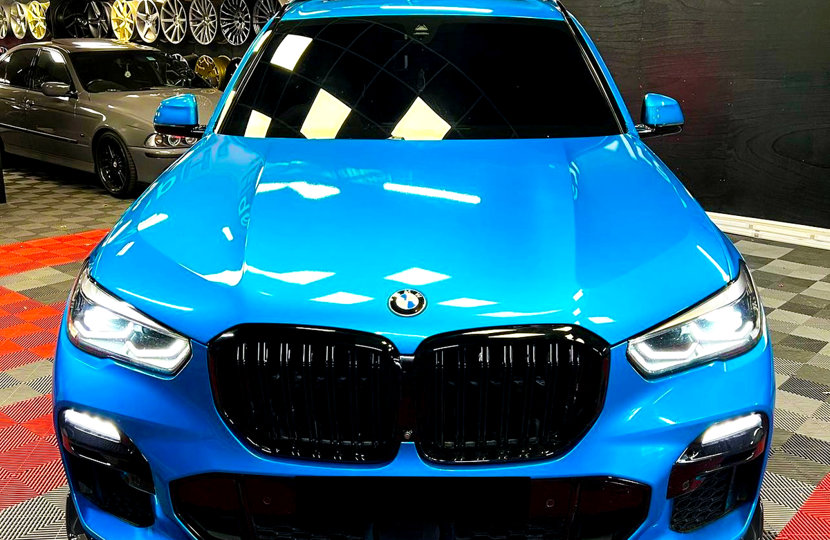 A blue BMW SUV is parked with headlights on in a showroom, surrounded by various wheels displayed on the wall and a checkered floor pattern.