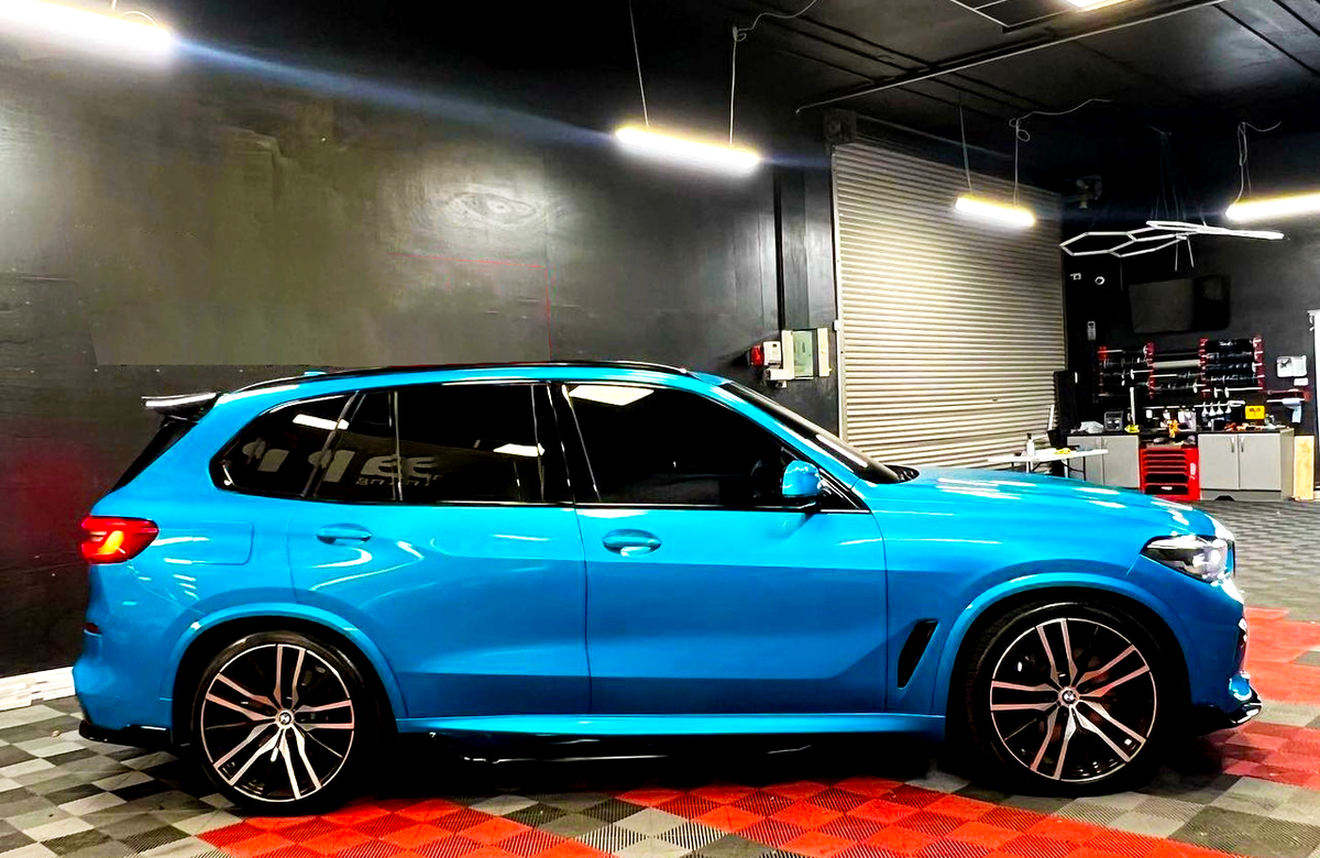 A bright blue SUV is parked in a garage with black walls and a checkered floor, under fluorescent lights, near a rolling door and shelves with tools.