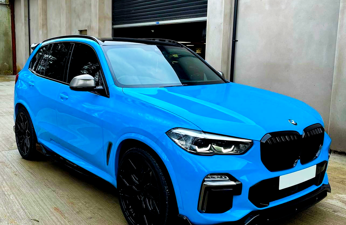 Bright blue SUV parked on a concrete surface, facing nearly forward. The car is in front of a tall, gray industrial building with a closed, dark roller door.