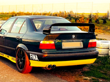 A modified black and yellow BMW car with a large rear spoiler is parked on a racetrack, surrounded by tires and a wire fence. Visible text includes 
