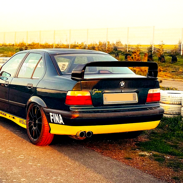 A modified black and yellow BMW car with a large rear spoiler is parked on a racetrack, surrounded by tires and a wire fence. Visible text includes "FINA," "MICHELIN," and "HS."