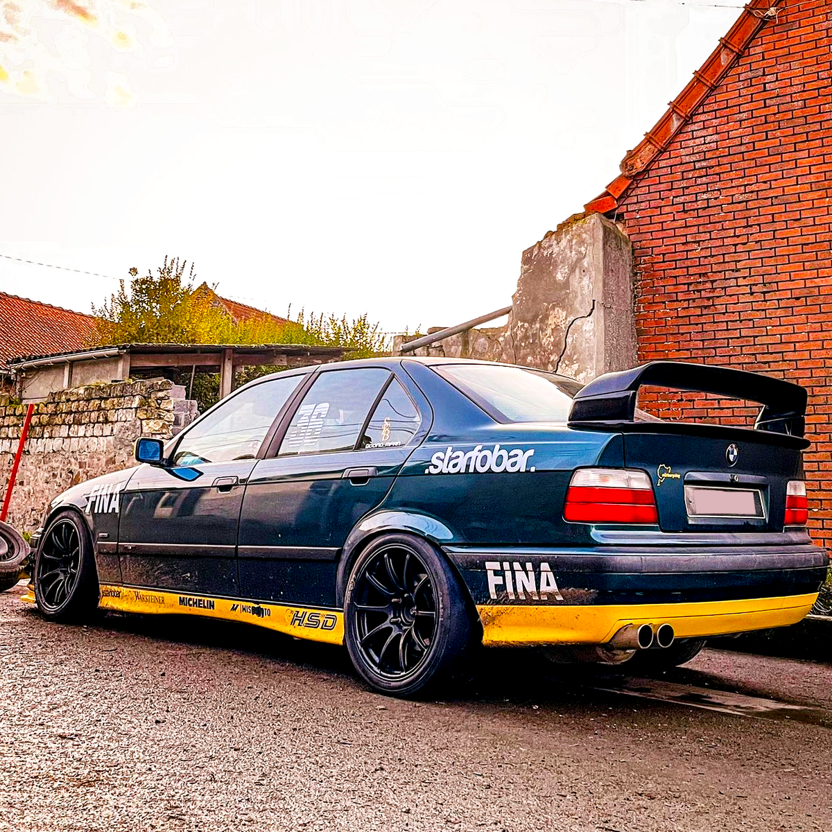 A modified blue car with a large rear spoiler and black wheels is parked on a street beside brick and stone walls. Text on the car reads "starobar," "FINA," "Michelin," and "HSD."