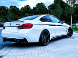 A white BMW 440i is parked on a paved street, against a backdrop of lush green trees and a cloudy sky.