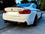 A white BMW M440i car is parked on a street, surrounded by trees under a clear blue sky.