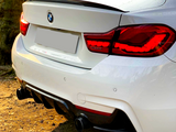 A white BMW car, parked on a dirt surface, has its red taillights illuminated. The rear bumper and dual exhausts are visible, with a shadow cast in the golden hour sunlight.