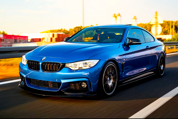 A blue BMW car speeds down a highway with blurred motion. The background shows industrial buildings and greenery under a clear sky.