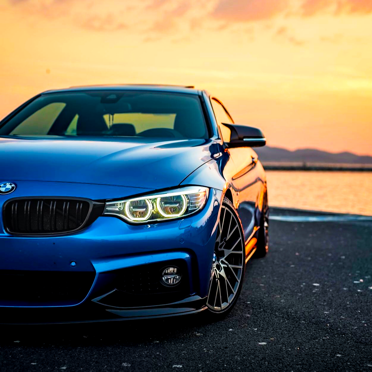 A blue car is parked by a waterfront at sunset, with its headlights on and mountains in the background.