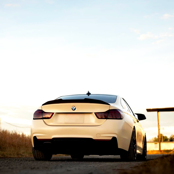 A white BMW car is parked on a dirt road during sunset, surrounded by open fields and a clear sky with a wooden structure in the background.