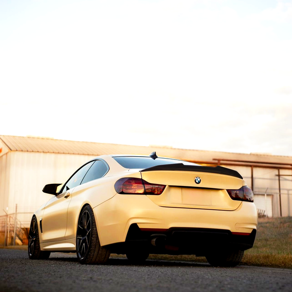 A yellow sports car is parked on a paved surface near a large, light-colored industrial building under a partly cloudy sky.