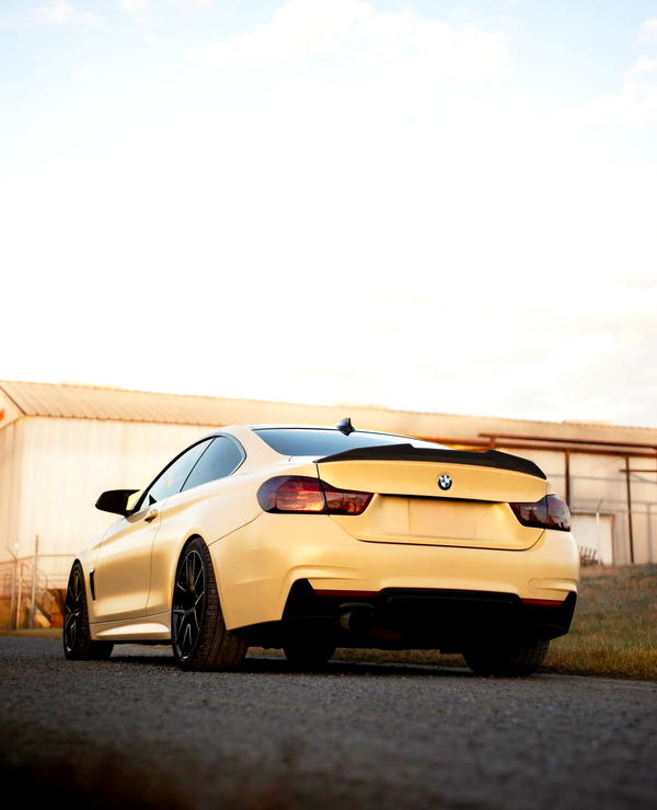 A yellow sports car is parked on a paved surface near a large, light-colored industrial building under a partly cloudy sky.