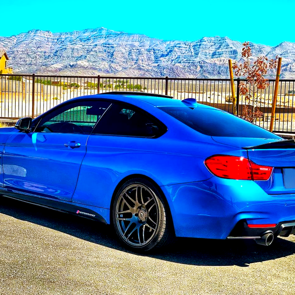 A blue sports car is parked on a sunlit road. It features dark wheels and a rear spoiler, with mountains and a clear sky in the background.