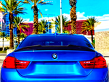 A blue BMW car is parked, facing a row of tall palm trees under a bright blue sky, with a modern building in the background.