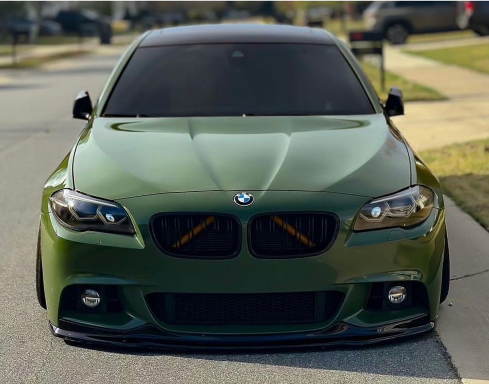 A green BMW car is parked on a suburban street, facing the viewer. Its tinted windows and sleek design emphasize a modern, stylish appearance. Lawns and driveways are visible in the background.