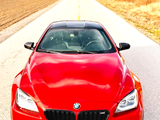 A red BMW car is parked on a rural road, surrounded by fields and trees, under a clear sky.