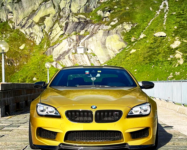 A yellow BMW sports car is parked on a stone road, with a mountainous landscape featuring green vegetation and rocky cliffs in the background.