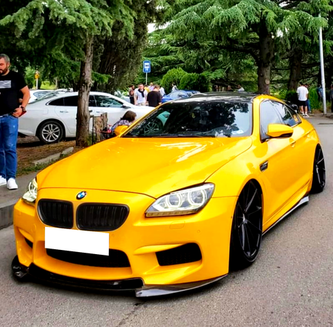 A yellow BMW sports car is parked on a street, surrounded by trees and people. Other parked cars and individuals conversing in the background create a lively atmosphere.
