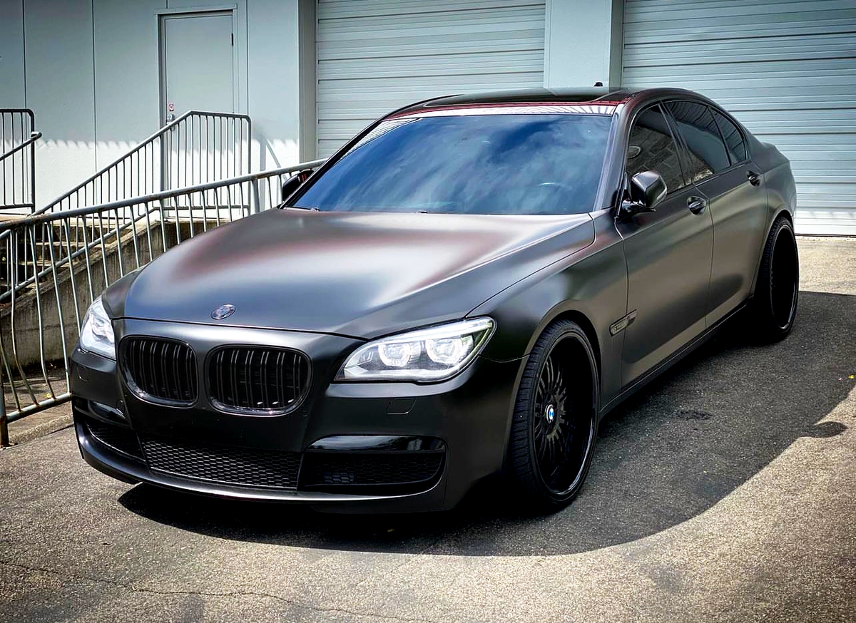 A sleek, matte black luxury sedan is parked on a paved surface. The vehicle is in front of a building with closed garage doors and a metal railing.