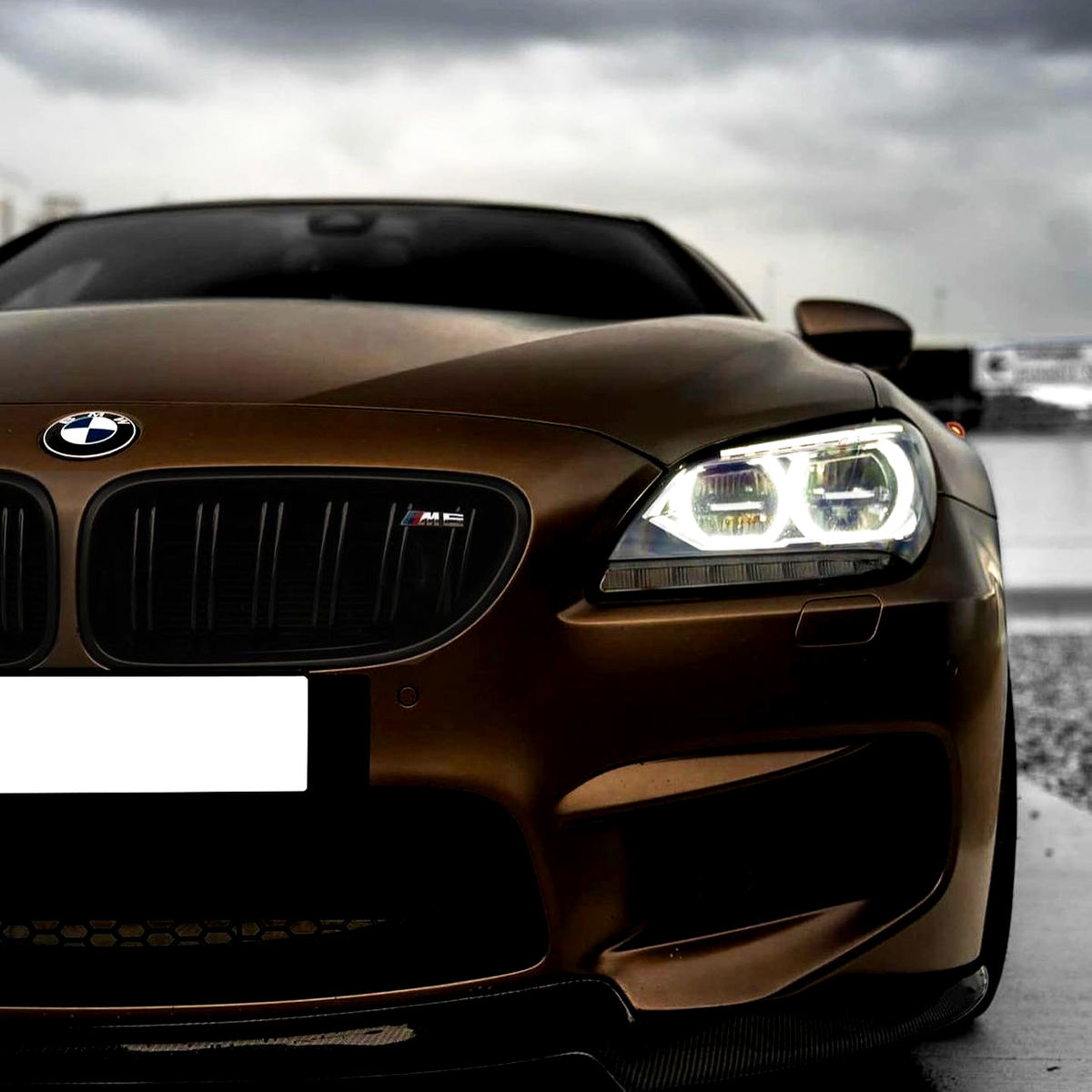 A matte brown BMW car with illuminated headlights, featuring a logo on the grille. It is parked on a wet surface under a cloudy sky.