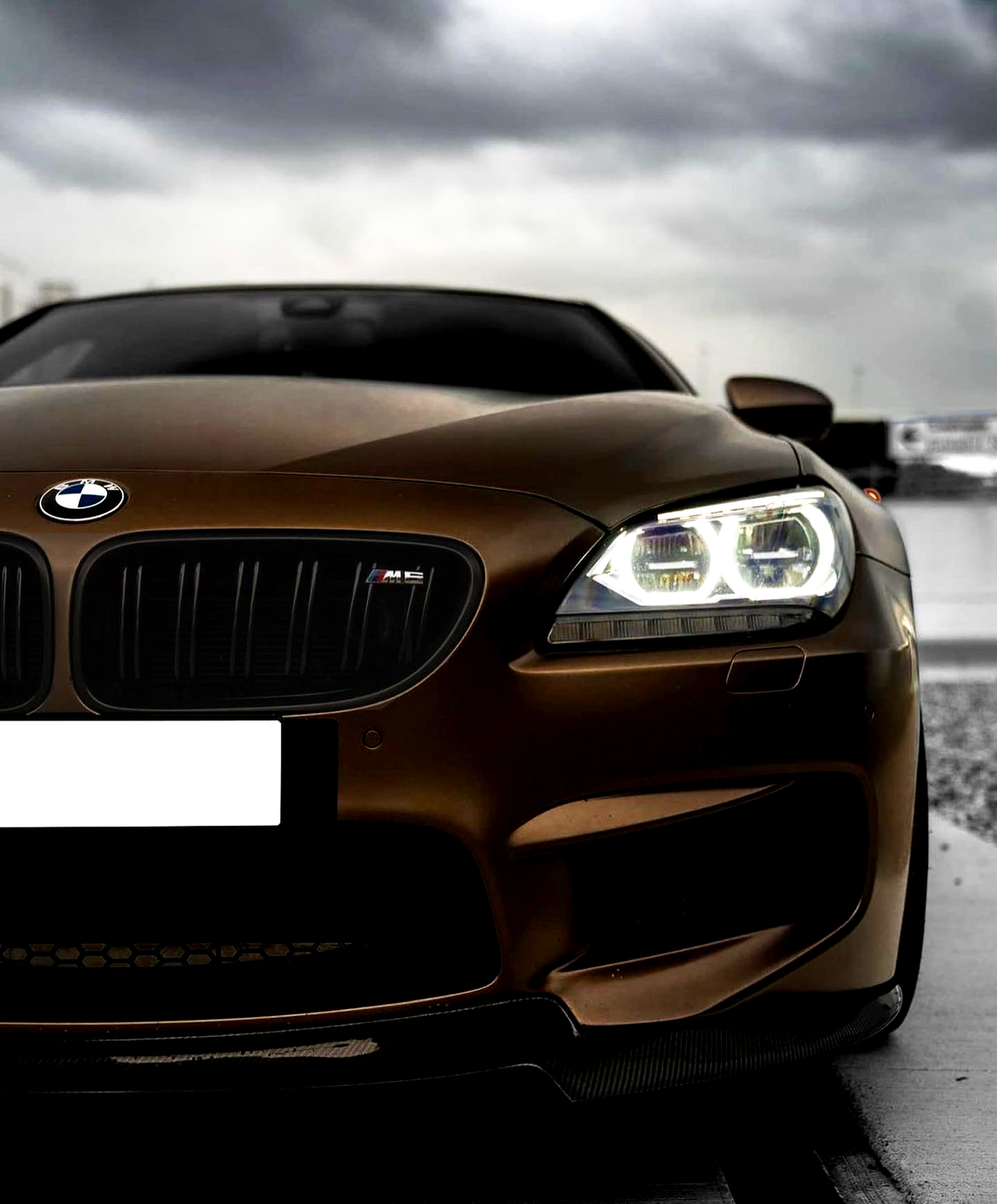 A matte brown BMW car with illuminated headlights, featuring a logo on the grille. It is parked on a wet surface under a cloudy sky.