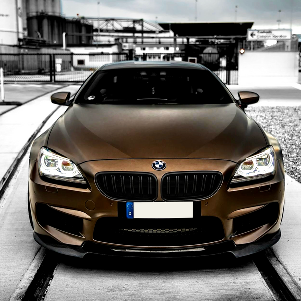 A brown BMW car, with headlights on, parked on a concrete surface in an industrial area. A sign in the background reads "Einfahrt Nordtor."