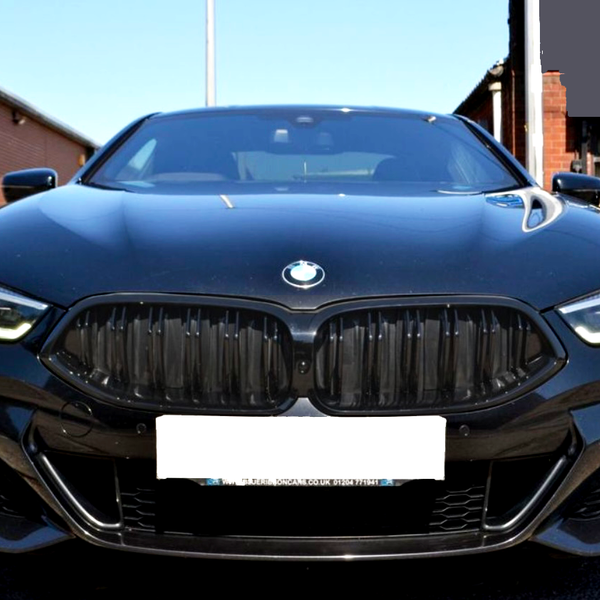 A sleek black car with illuminated headlights is parked on an asphalt surface between brick buildings. The BMW emblem is visible, and the license plate reads "WWW.TDREID.CO.UK 01224 871 531".