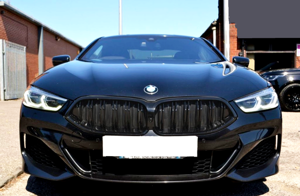 A black BMW car is parked outdoors, facing forward. Its LED headlights are illuminated. Behind it are brick buildings and another black car. The license plate reads: "STJAMESCARS.CO.UK 01244717163."
