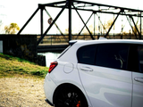 A white car, parked on a gravel road, is in the foreground, with a metal bridge in the background. Bright sunlight casts distinct shadows, highlighting the car's red brake calipers.