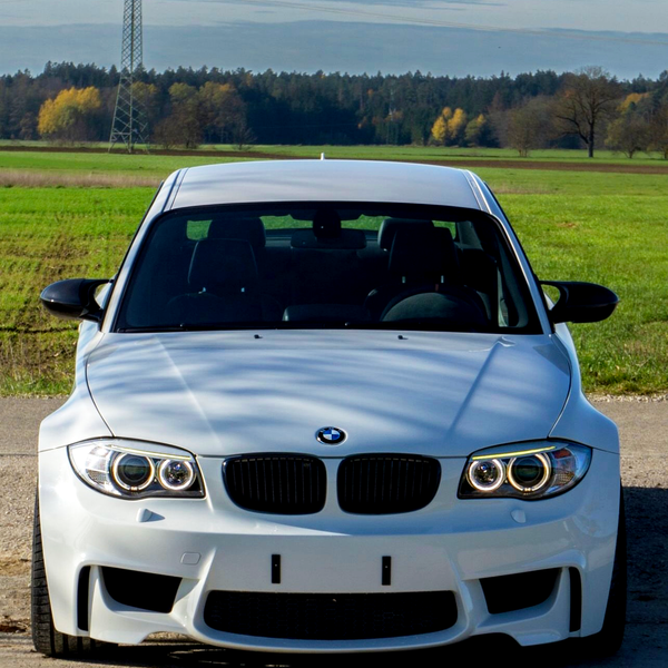 A white BMW car is parked on a rural road. It is facing forward with a power line in the background and surrounded by green fields and distant trees.