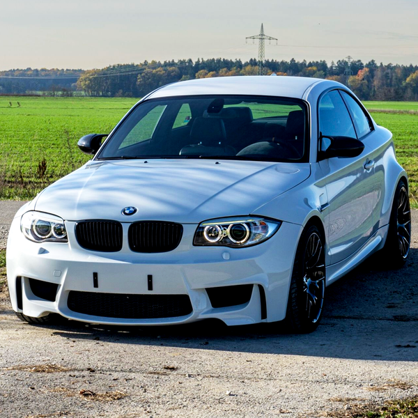 A white sports car is parked on a rural road with green fields and trees in the background under a clear sky.