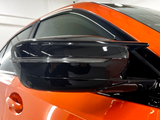 A sleek, black side mirror on an orange car is shown indoors, with reflections on its glossy surface, under bright lighting.