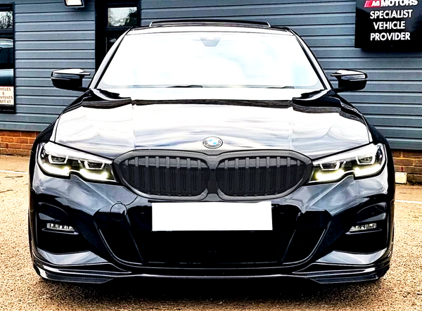 A sleek black BMW car, headlights on, facing forward, is parked in front of a gray paneled building. Nearby signage reads: "A6 MOTORS SPECIALIST VEHICLE PROVIDER."