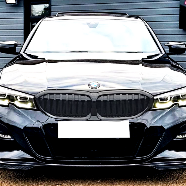 A black BMW car faces forward, parked on a light gravel surface, with a sign reading "A6 MOTORS SPECIALIST VEHICLE PROVIDER" on the building in the background.