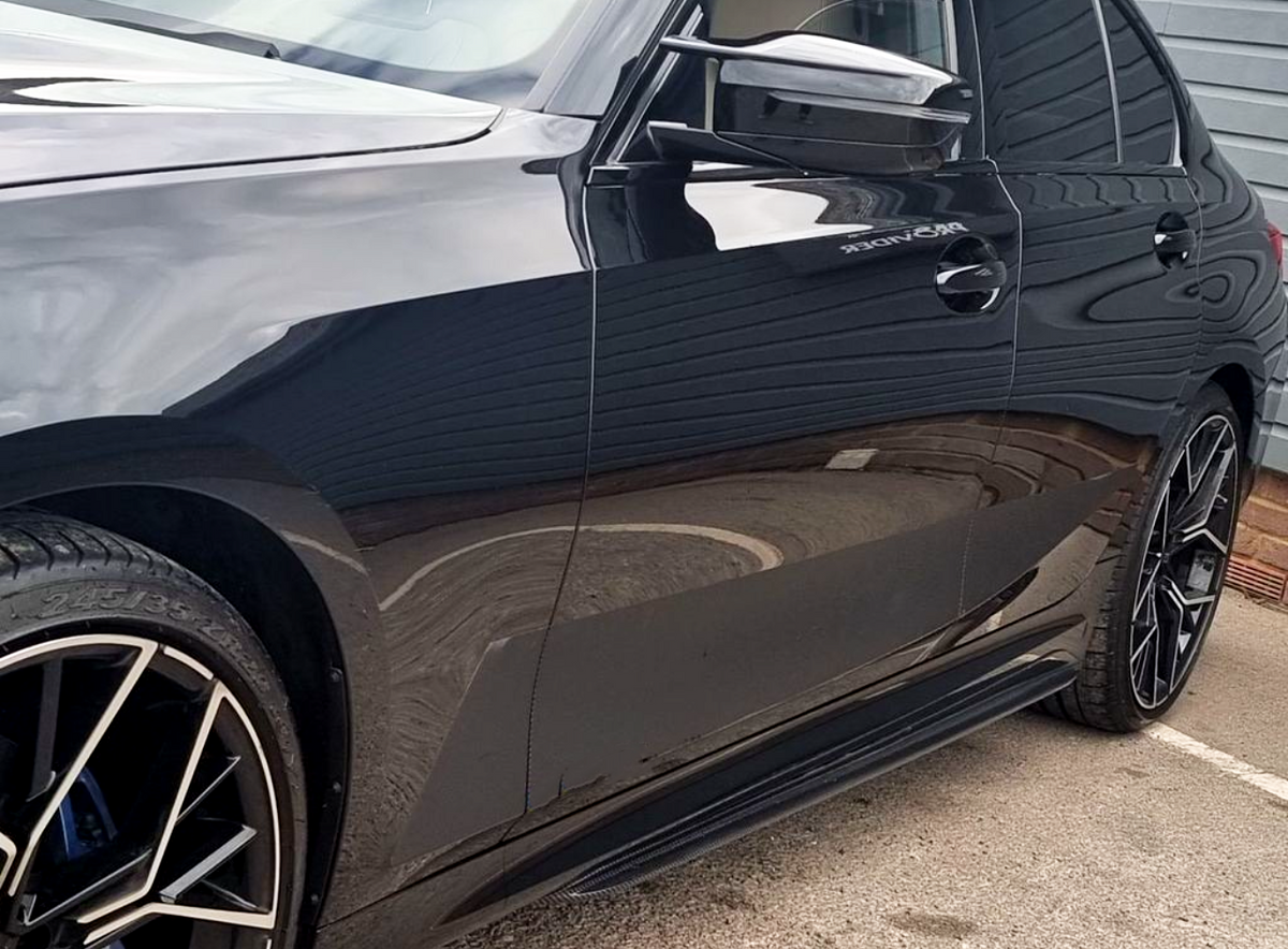 A sleek, black car is parked, reflecting its surroundings. Visible elements include the front tire and side mirror, with partial text "BRONDER" near the door. It rests on concrete.