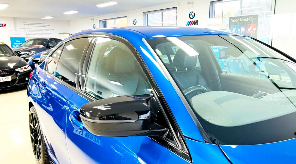 A shiny blue car is parked in a well-lit showroom, surrounded by other vehicles. Visible text includes “Your Next Car” and “MotoNovo,” indicating a promotional setting.