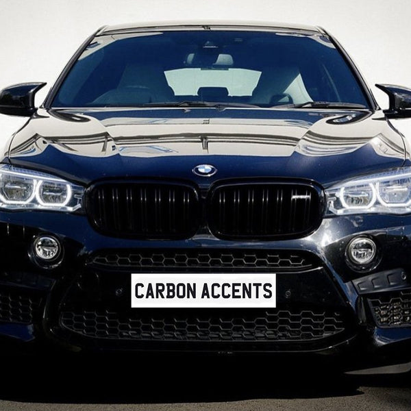 Car with black grille, shining headlights, and reflective surface, parked on asphalt against a plain white wall. License plate displays text: "CARBON ACCENTS."