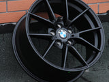 A black BMW wheel rim leans against a textured wall with visible metallic spokes and the BMW logo at the center. The background features orange brickwork and a gray surface.