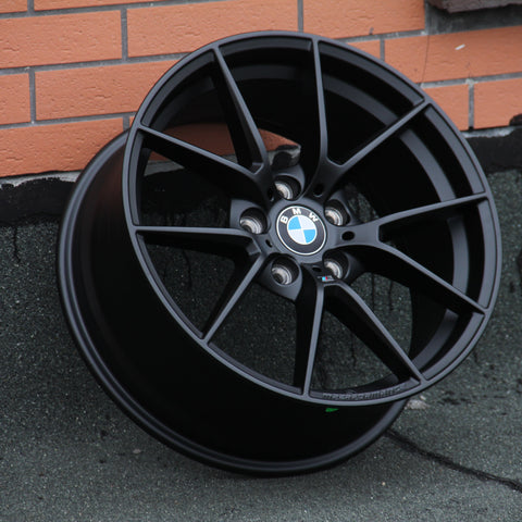 A black BMW wheel rim leans against a textured wall with visible metallic spokes and the BMW logo at the center. The background features orange brickwork and a gray surface.