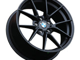 A black BMW alloy wheel with a multi-spoke design is positioned against a white background. The BMW logo is centered, surrounded by lug nut holes. 
