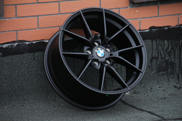 A black BMW wheel leans against an orange brick wall on a textured gray surface. The BMW logo is centrally visible.