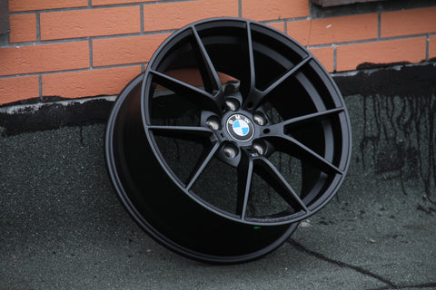 A black BMW wheel leans against an orange brick wall on a textured gray surface. The BMW logo is centrally visible.