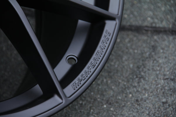 A matte black car wheel rim embossed with "M PERFORMANCE" is positioned on a textured, dark surface. A bolt hole is visible, adding detail to the sleek design.