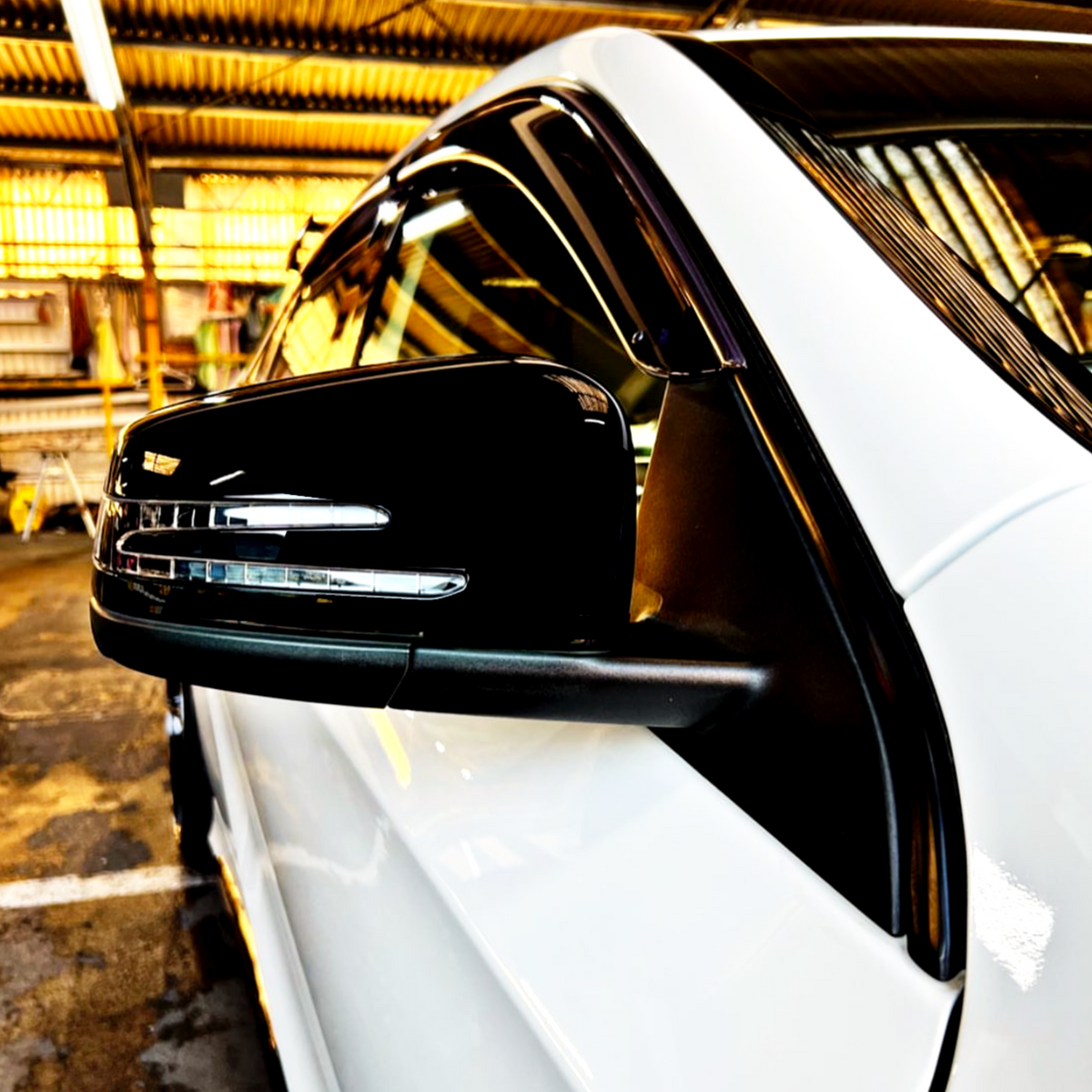 A car's side mirror with LED indicators is prominently displayed, reflecting light. The vehicle, white with glossy black trim, is parked inside an industrial garage setting.