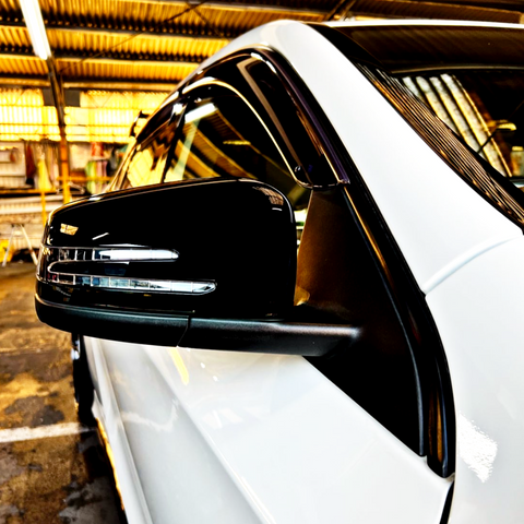 A black side mirror on a white car is parked in a well-lit garage, surrounded by industrial equipment and yellow lighting.