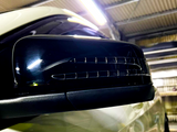 A close-up of a shiny black car side mirror, reflecting light, inside a large, well-lit garage with a corrugated metal ceiling.