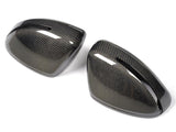Two carbon fiber car side mirror covers rest on a white surface, displaying a glossy, woven texture with elongated openings near the edges, reflecting light.