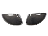 Two carbon fiber wing mirror covers are side by side, angled towards each other, against a plain white background. The covers have a glossy finish with a detailed woven pattern.